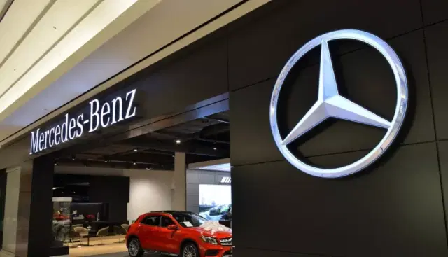 A Mercedes Benz car dealership showroom shows a partial view of a red hatchback car. A large Mercedes logo occupies the store exterior.